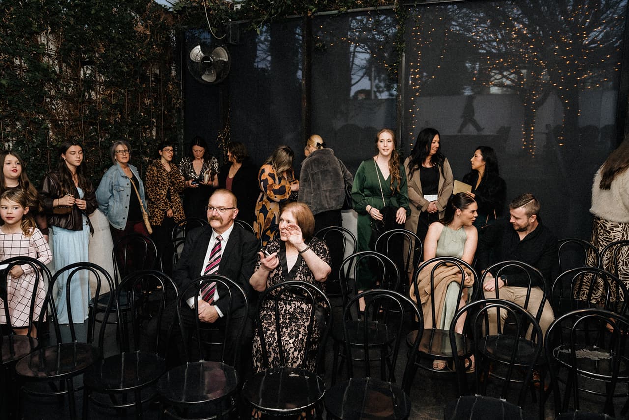 Guests finding their seats on black bentwood chairs in the covered courtyard, fairy lights and green walls behind them
