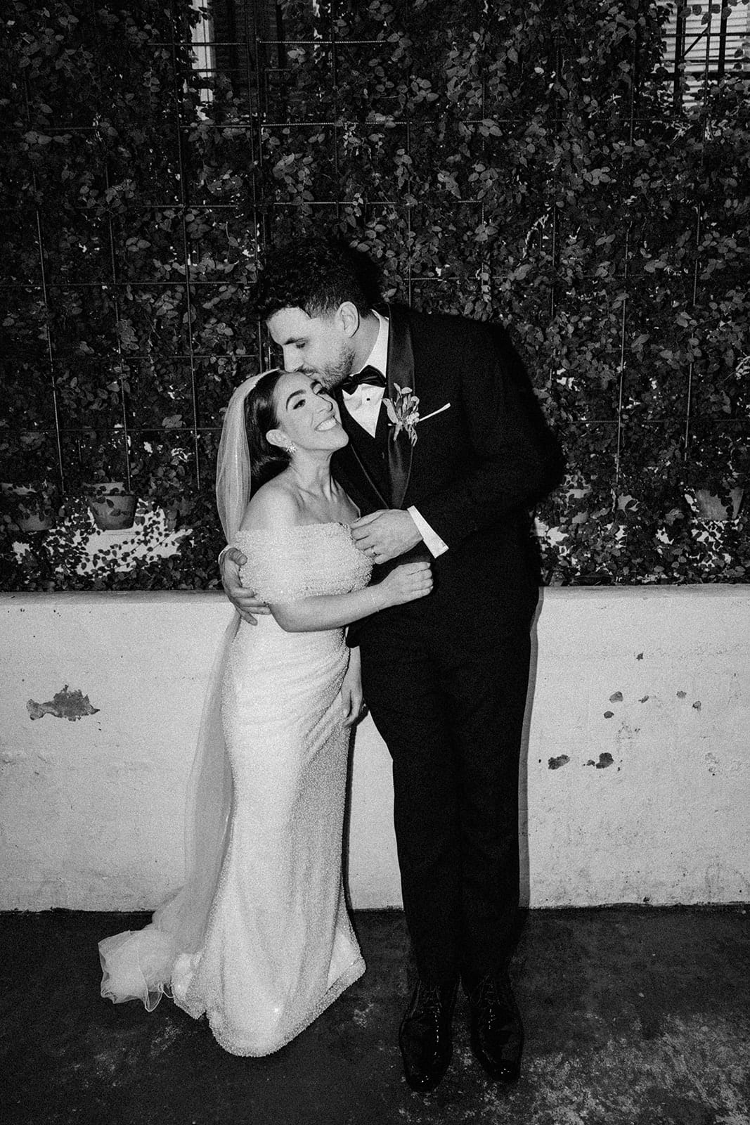 Black and white photo of George kissing Tia in front of the garden wall and the white brick planter