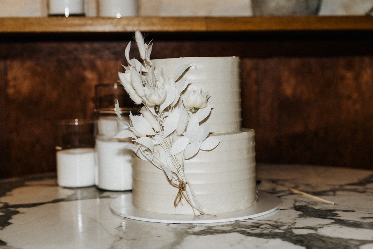 The white cake decorated with dried white flowers on a marble table, candles beside it