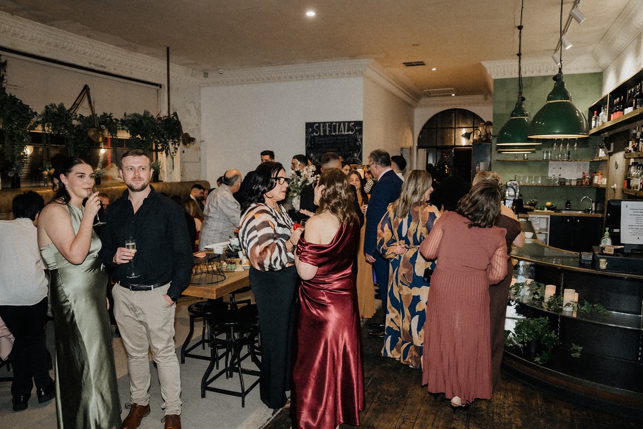 Guests mingling in the dining room under green pendant lamps, a chalkboard and an arched window beyond the bar