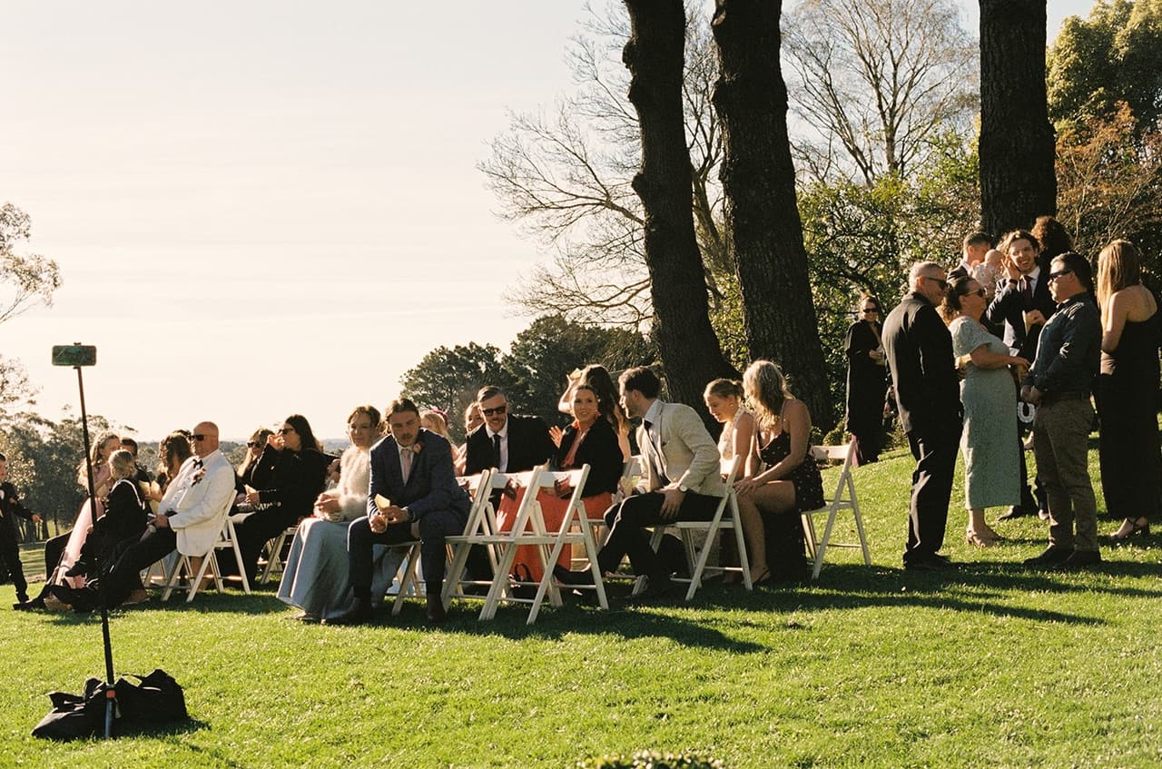 Guests settling into white folding chairs on the sloping lawn under the big trees, a phone on a stand in the foreground, on 35mm film