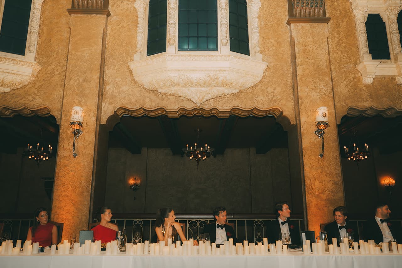 The bridal table behind rows of candles, beneath the ballroom’s arches, balconies and wall lamps