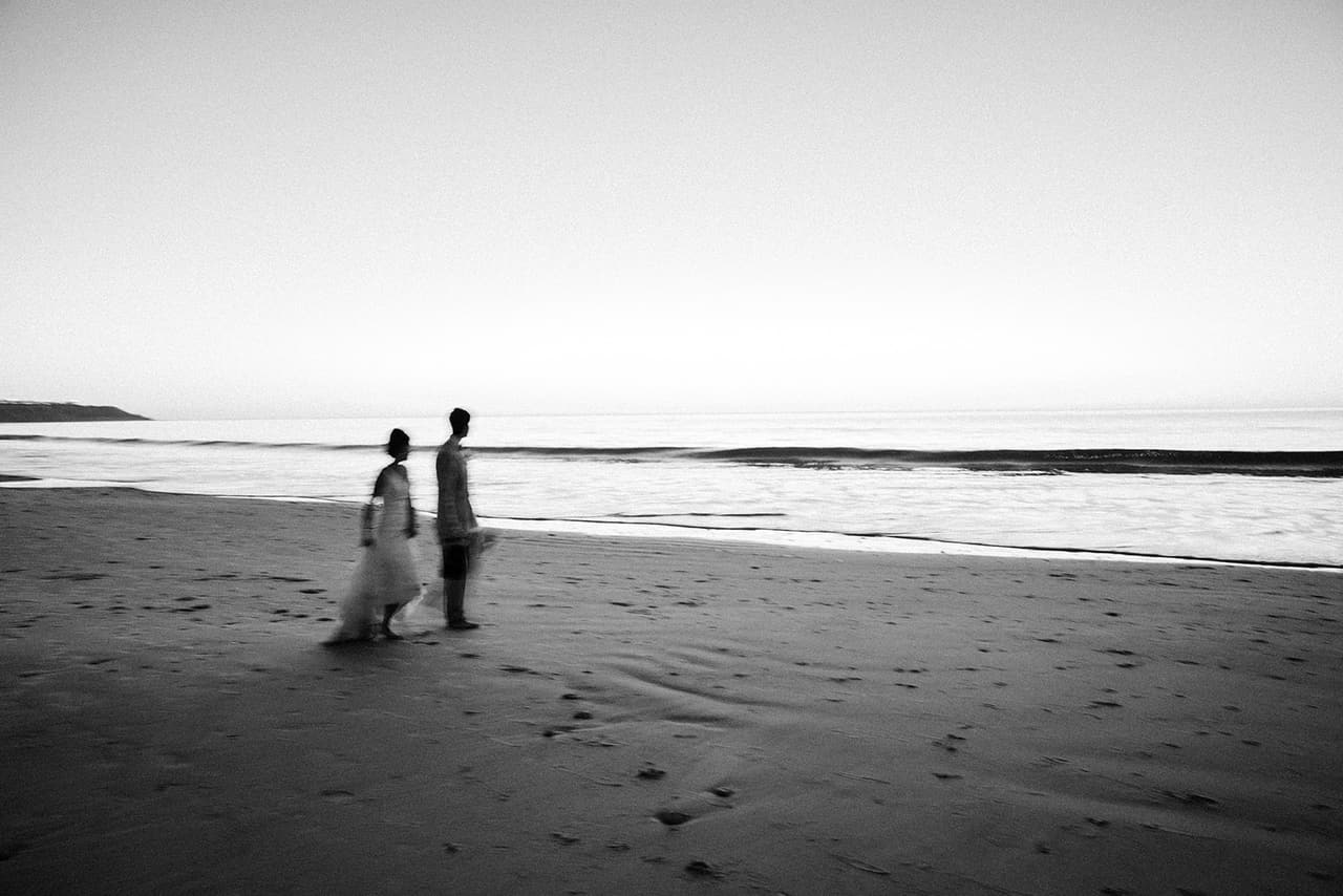 Black and white photo of the beach at dusk, Sayuri and Shuto standing at the water’s edge