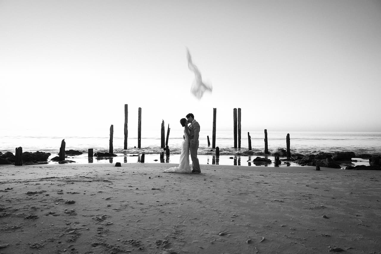 Black and white photo of the open sand in front of the old jetty posts, Sayuri and Shuto kissing as her veil flies above them