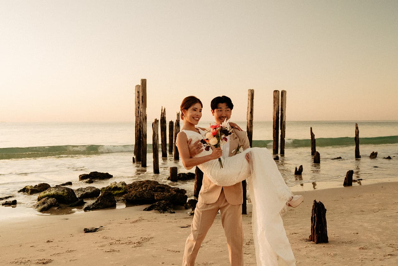 Shuto carrying Sayuri past the rocks and the old jetty posts at the water’s edge