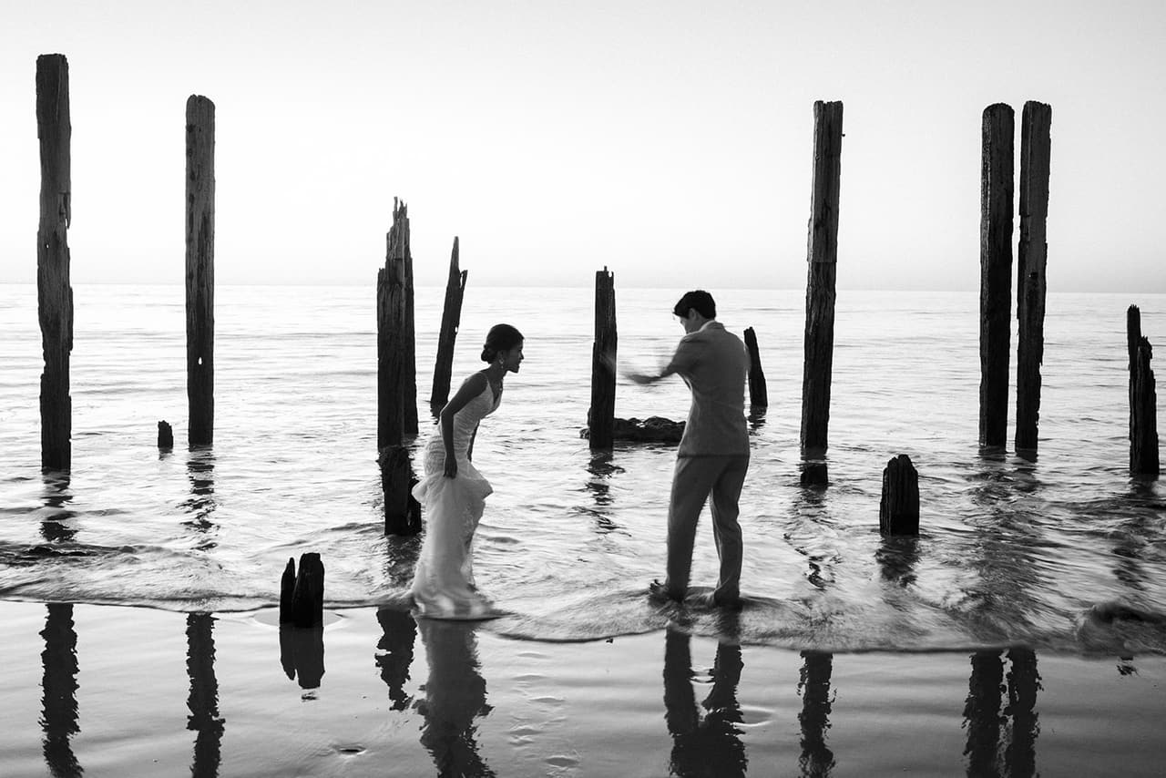 Black and white photo of Sayuri and Shuto wading in the shallows between the old jetty posts