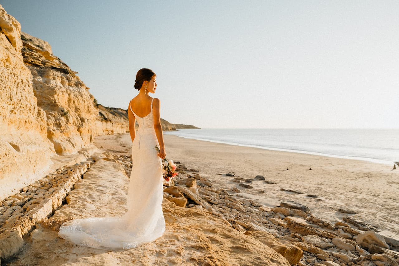 Sayuri seen from behind on the rock shelf below the cliffs, her train spread over the stone and the beach beyond