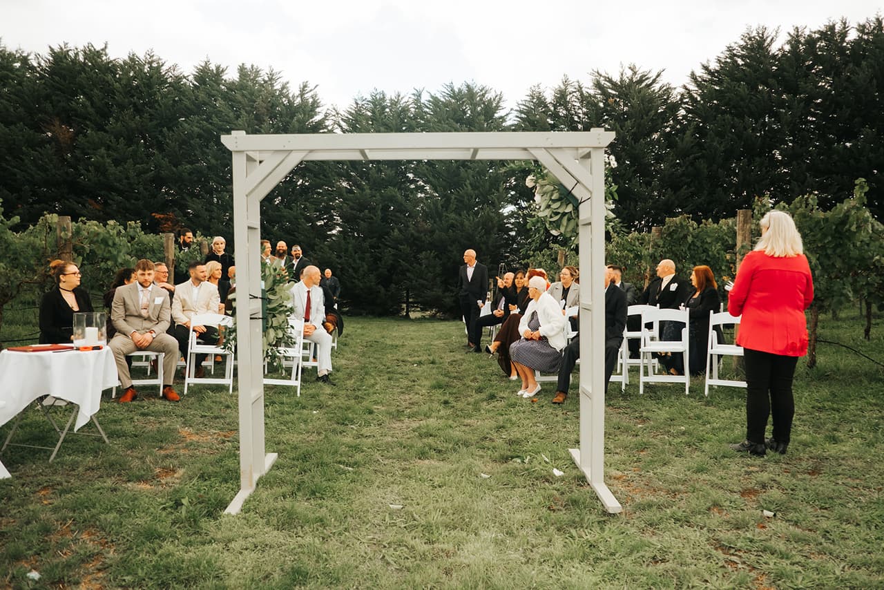 The white timber arbour on the grass between the vines, white folding chairs filling with guests