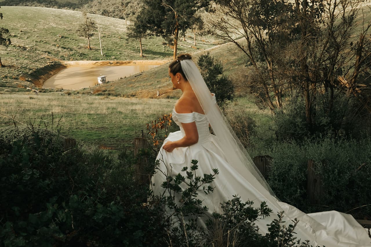 Jessica in her gown among the shrubs of the hillside garden, a farm dam and paddocks below