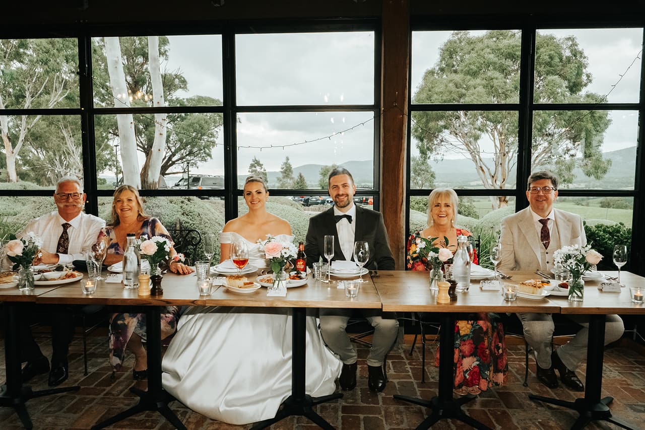 Jessica and Max at the head table with their family, tall windows and gum trees behind them