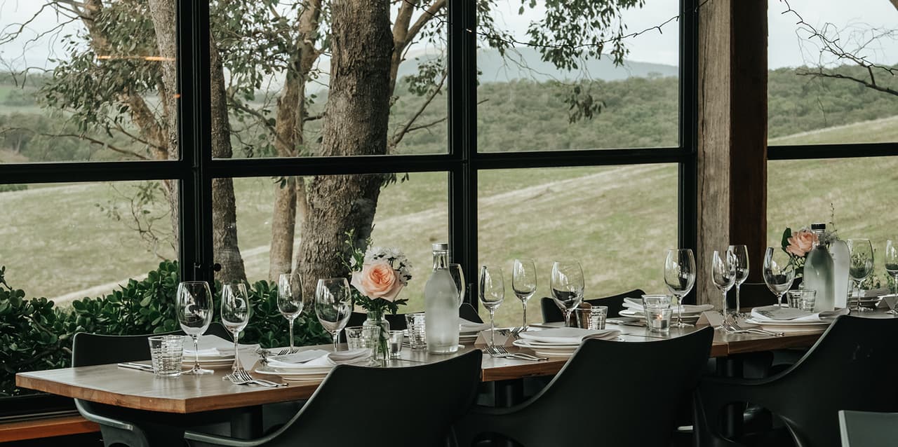A dinner table with glasses and pink roses beside the tall windows, gum trees and paddocks outside
