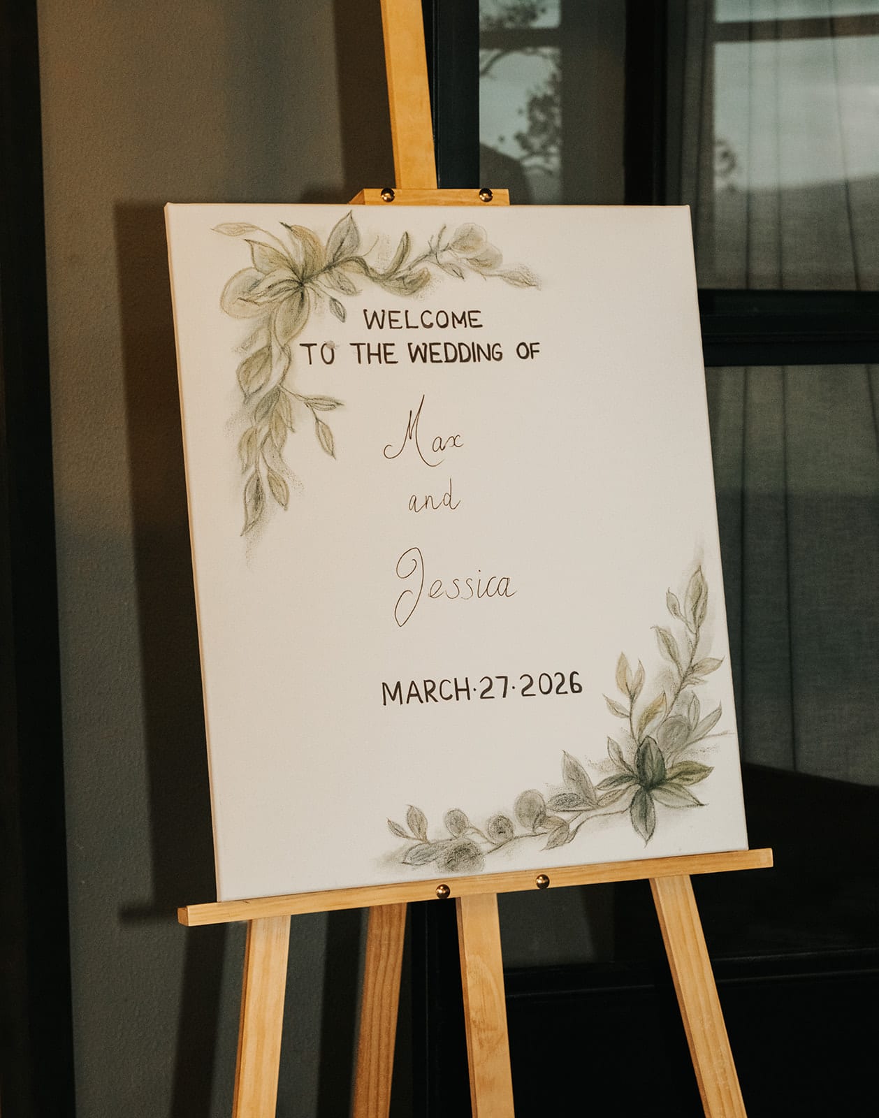 A welcome sign with Max and Jessica’s first names and the date, painted with green leaves, on a timber easel
