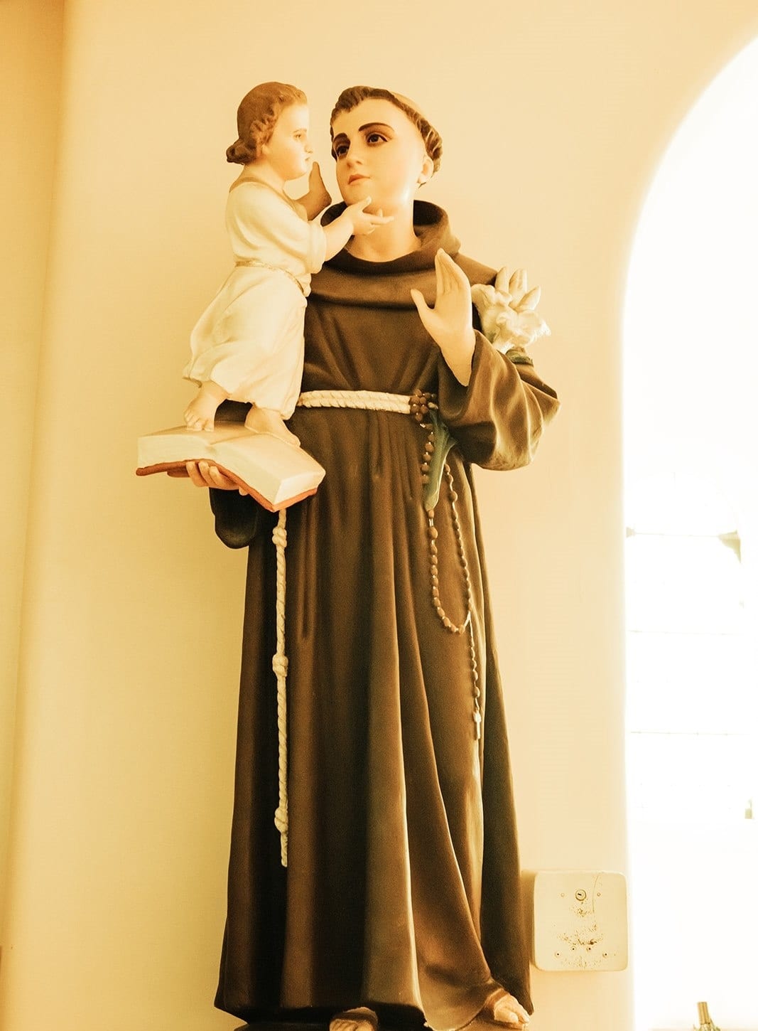 A statue of a friar in a brown habit holding the Christ child against a pale wall