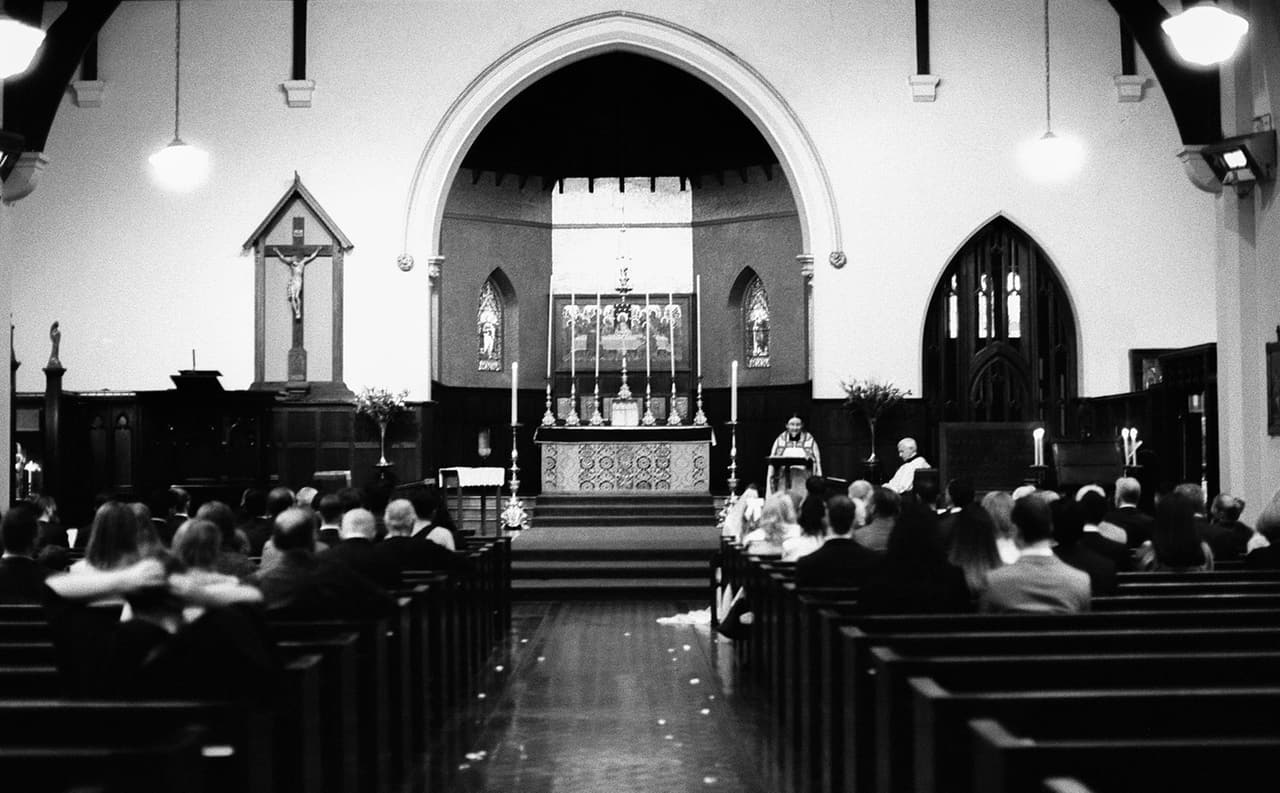 Black and white photo of the ceremony from the back of the church, guests filling the pews up to the arch and the altar, on 35mm film