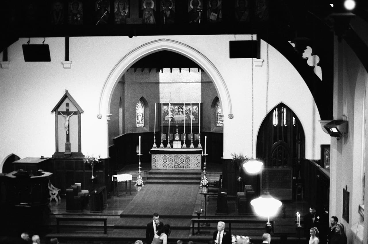 Black and white photo of the inside of St Peter’s seen from the gallery, the timber pews, a crucifix and the tall arch to the sanctuary and altar, on 35mm film
