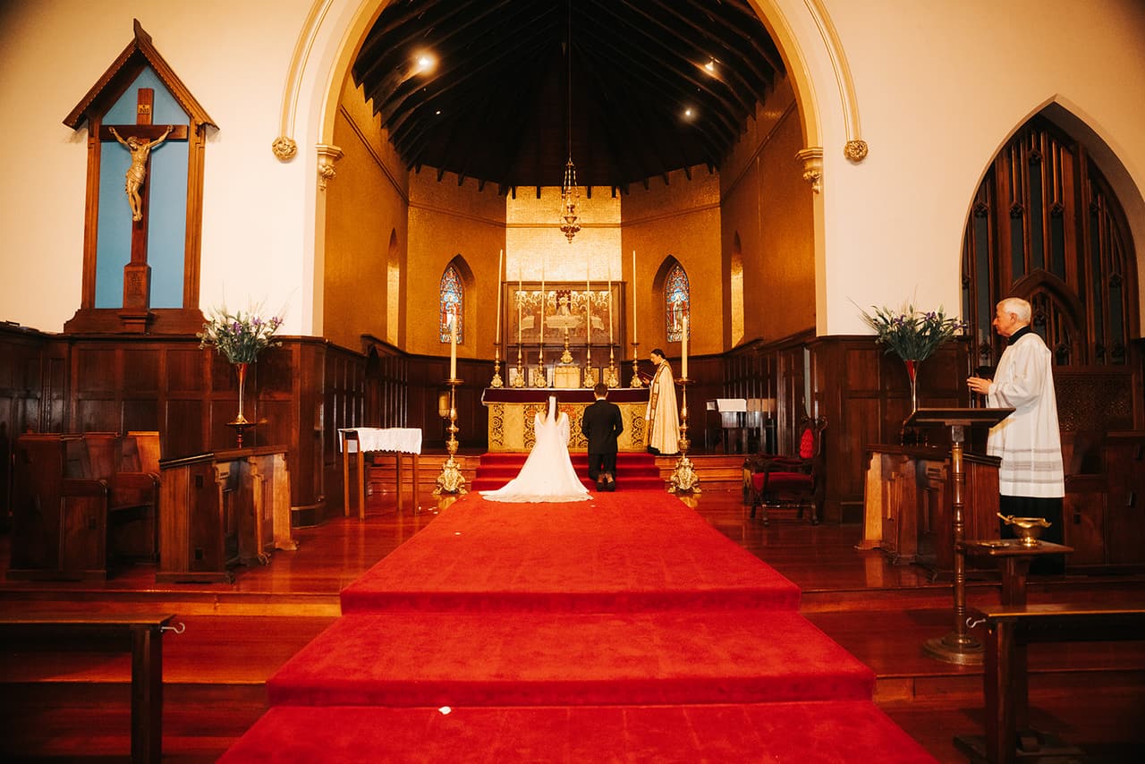 The red carpeted steps up to the high altar and its gold reredos, Bianca and Aidan kneeling at the top and a crucifix on the wall beside the arch