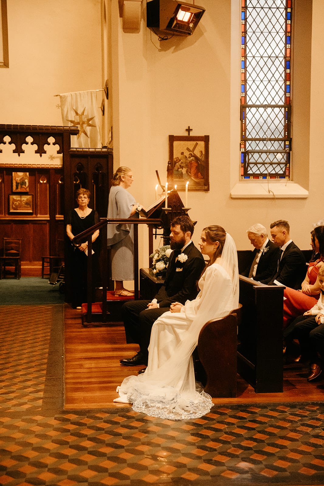 Bianca and Aidan seated at the front of the church beside a tall stained glass window, guests in the pews around them