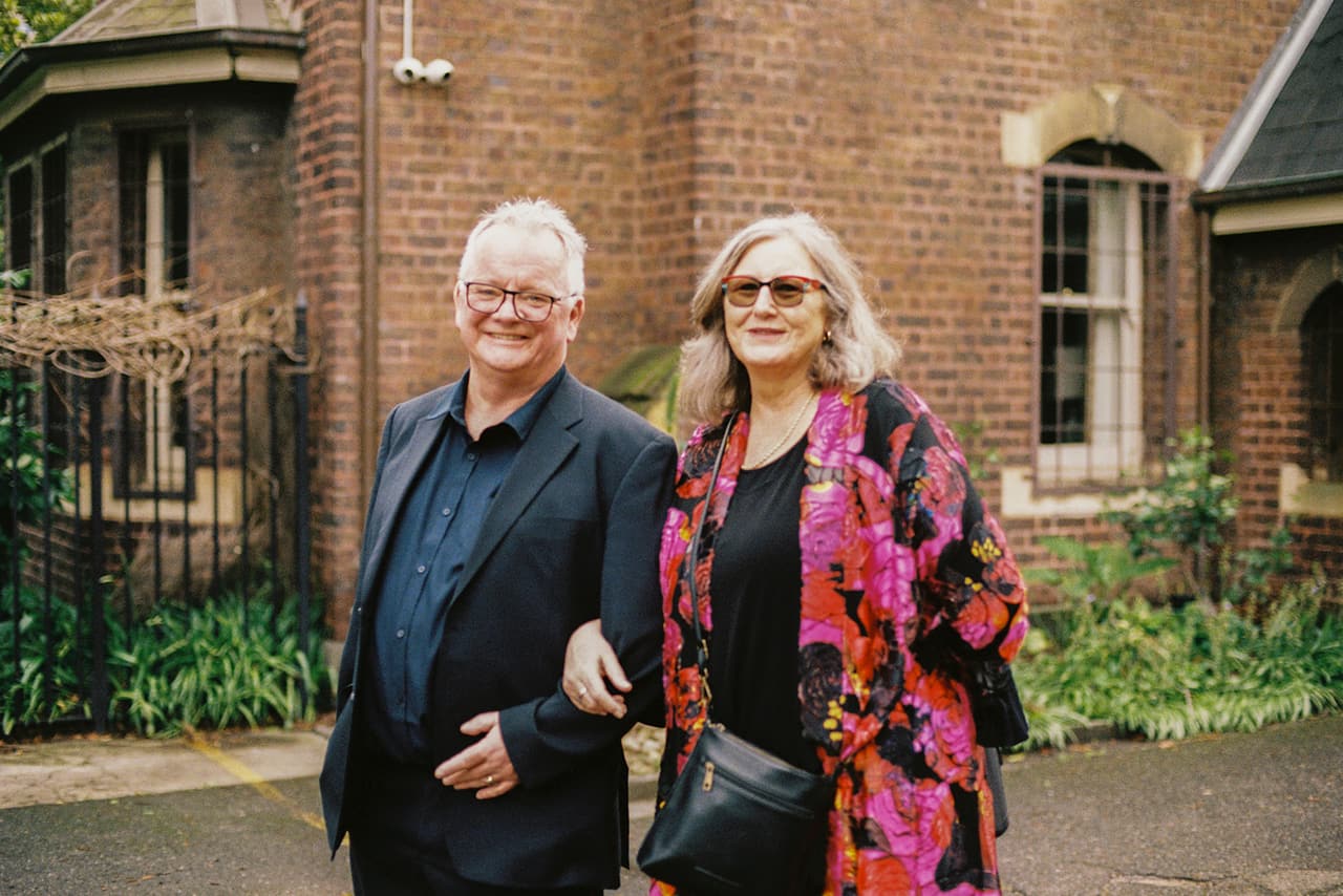 Two guests arm in arm in the church grounds in front of a red brick building, on 35mm film