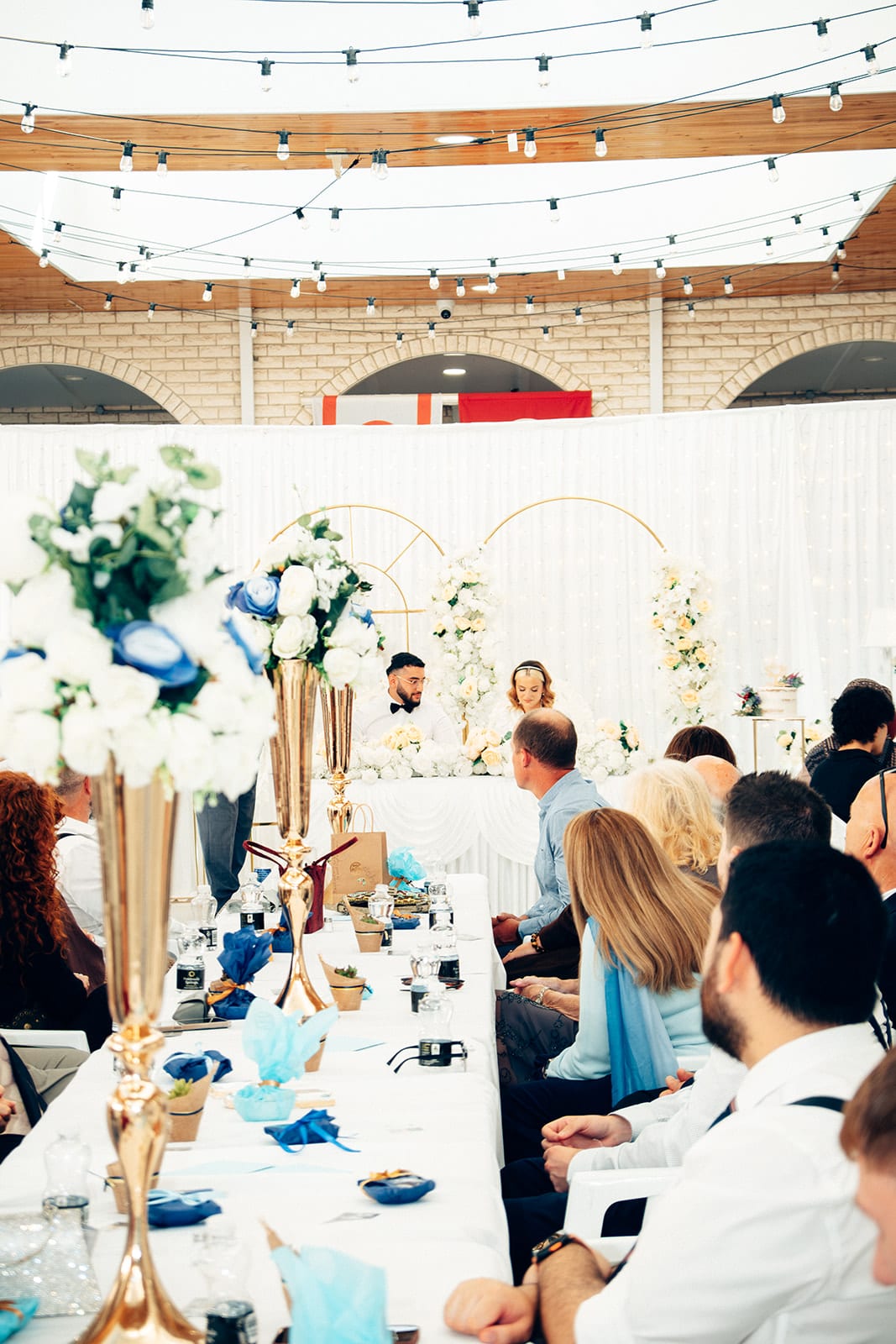 The view from behind the guests’ tables to the head table and its gold arches, white and blue flowers in tall gold vases along the tables
