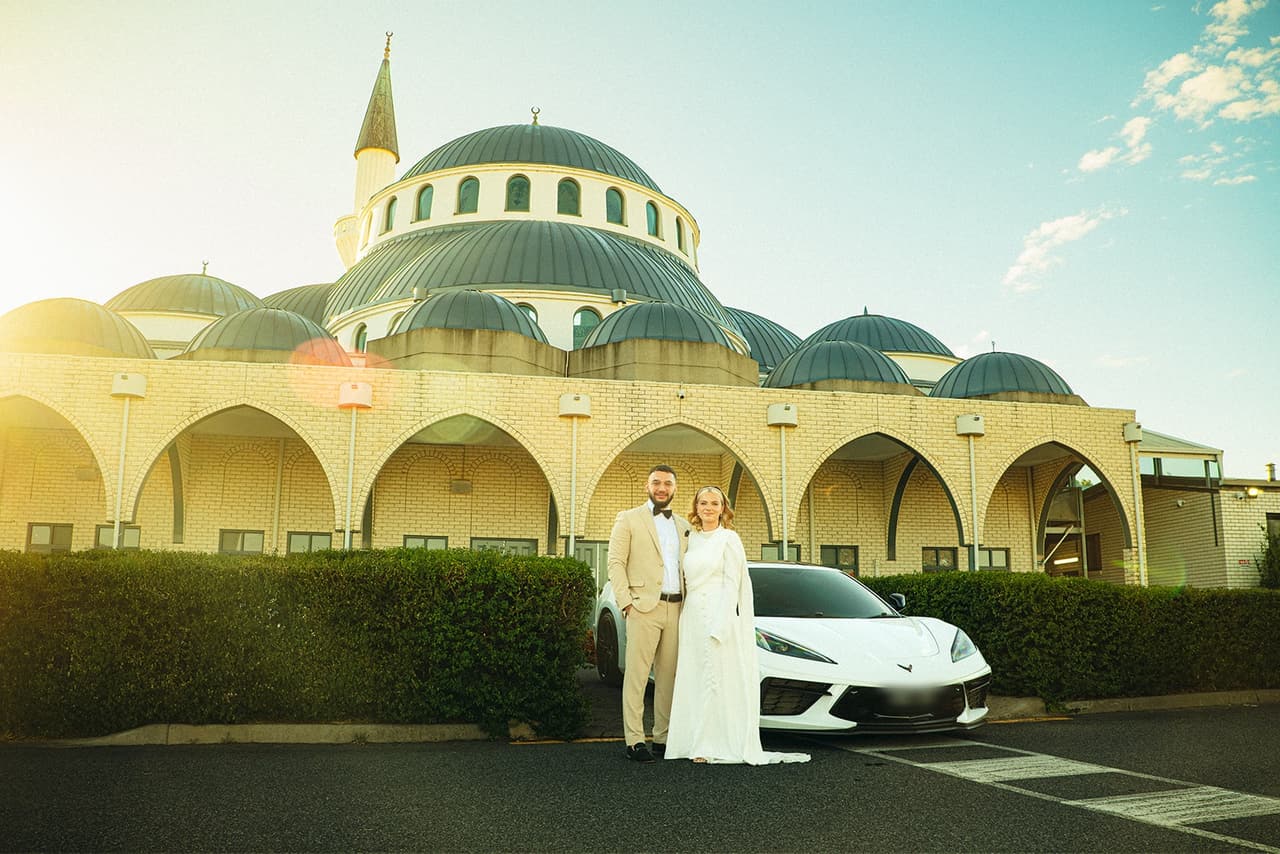 Elektra and Yasir beside a white sports car below the domes, minaret and arched front of the Sunshine Mosque in the late sun