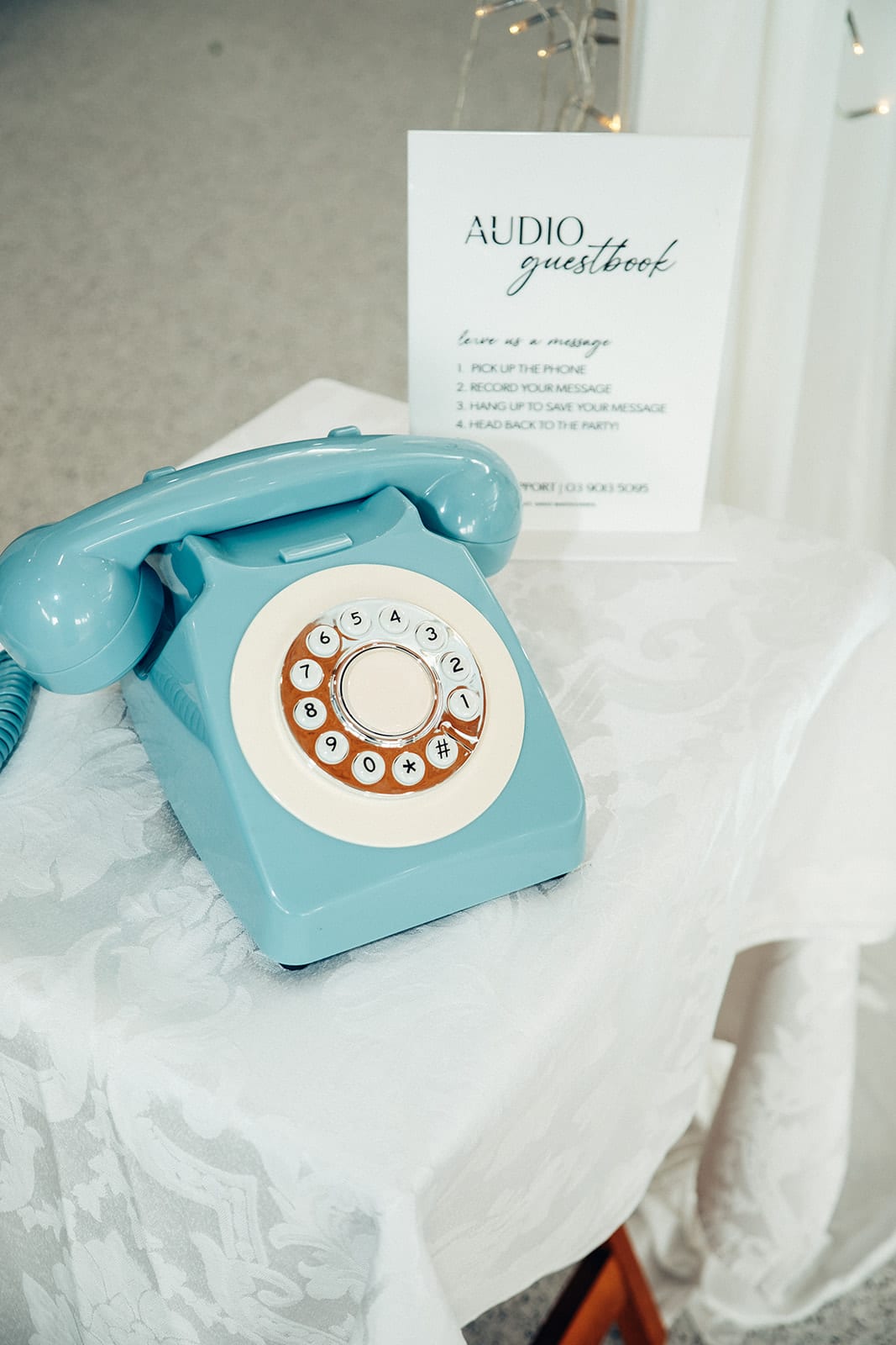 A blue rotary telephone for an audio guestbook on a lace cloth, its instructions on a card behind it