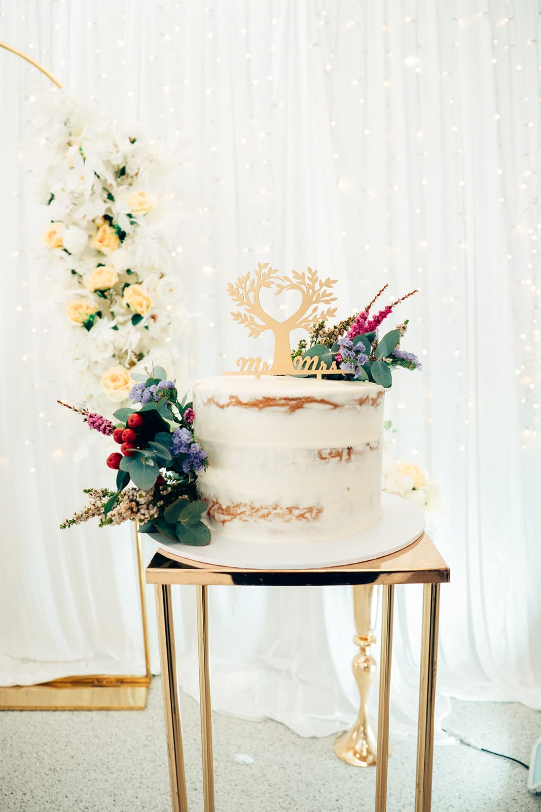 A semi-naked white cake with a gold heart tree and Mr and Mrs topper and fresh flowers, on a gold stand
