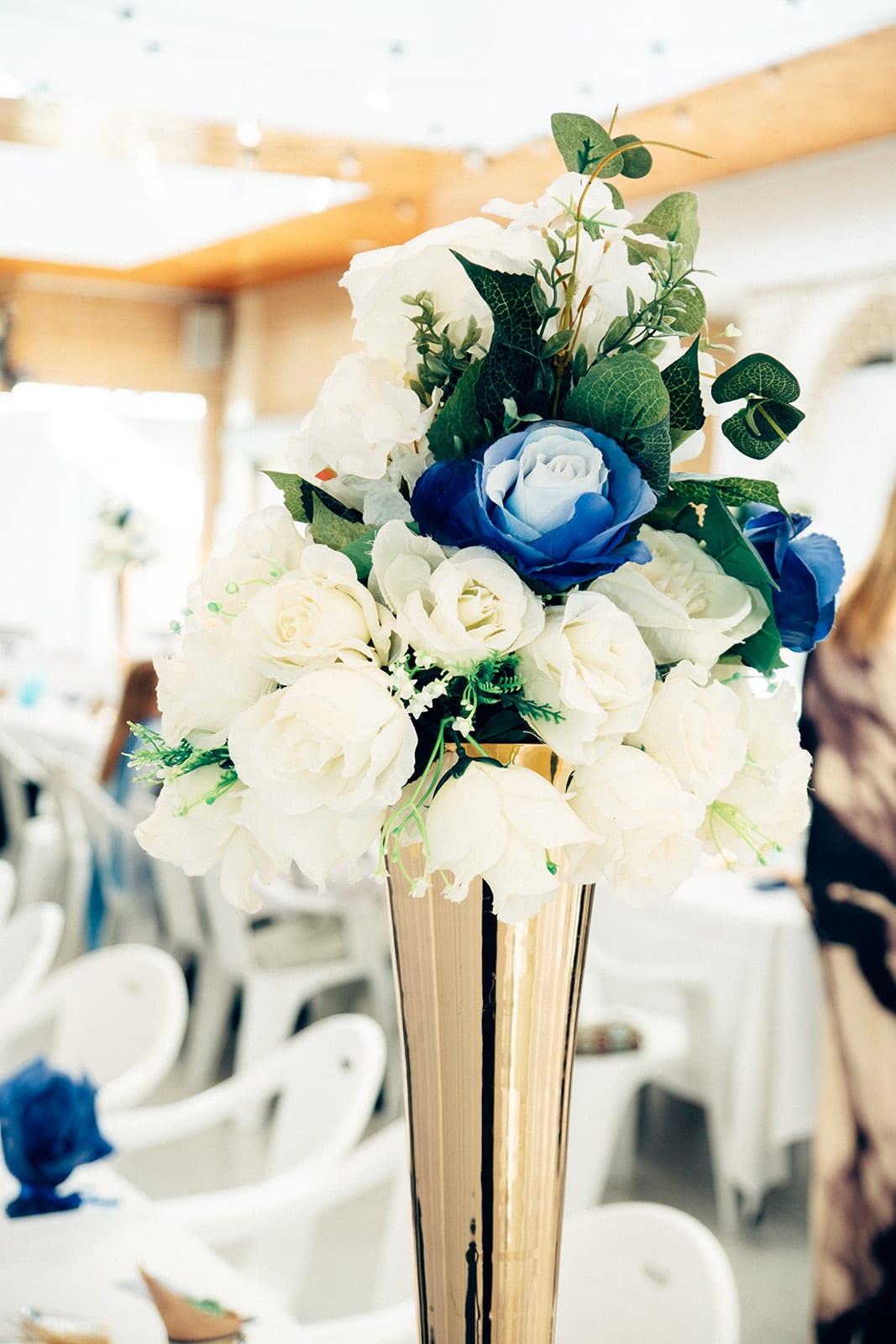 A tall gold vase of white and blue roses on a guest table, white chairs around it