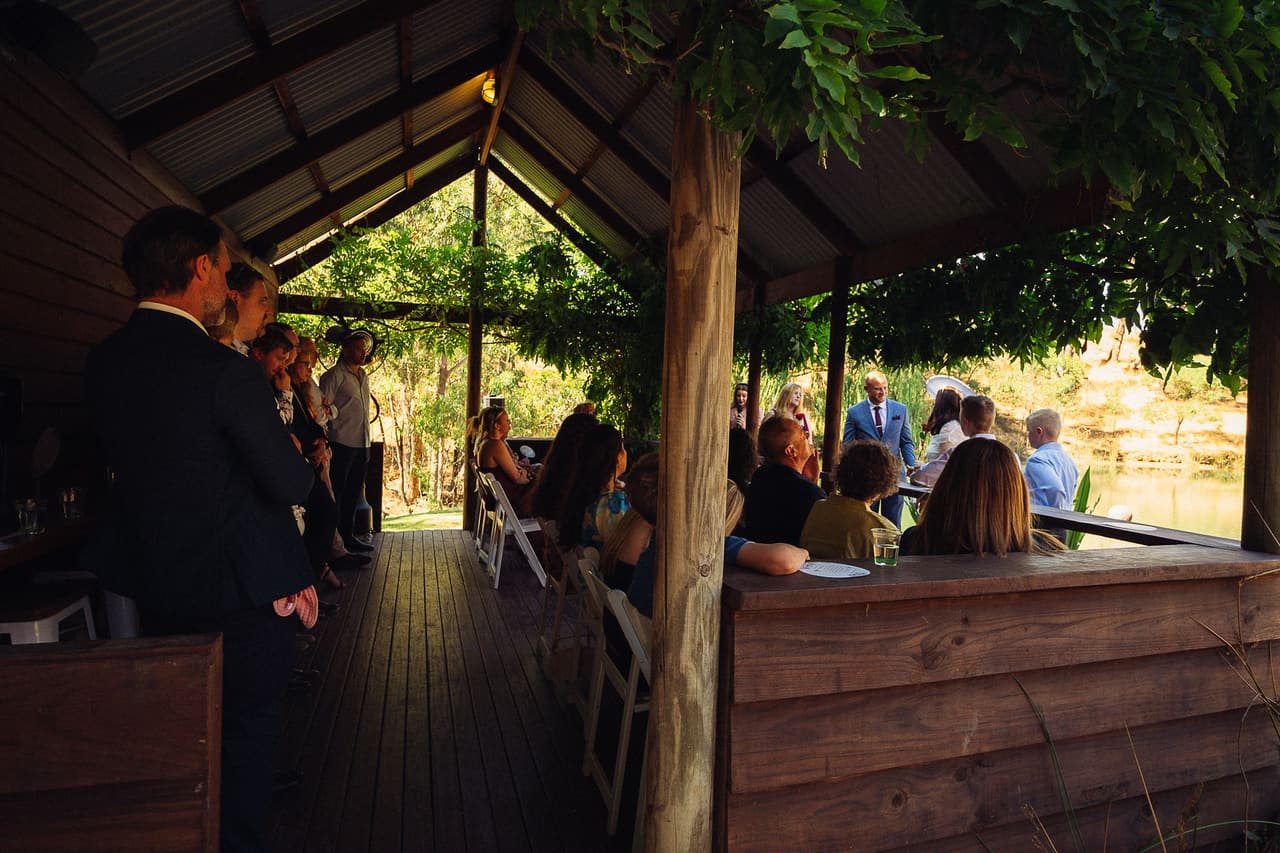 Inside the Boatshed during the ceremony, guests seated along the timber rail under the iron roof and the couple at the far end over the water