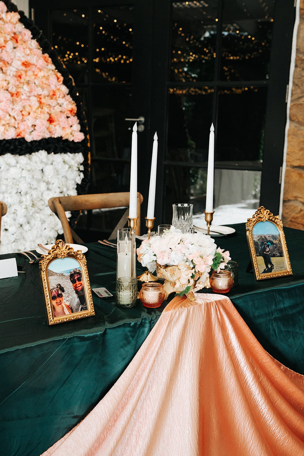 The head table with candles, a pink tablecloth and framed photos in front of the flower wall