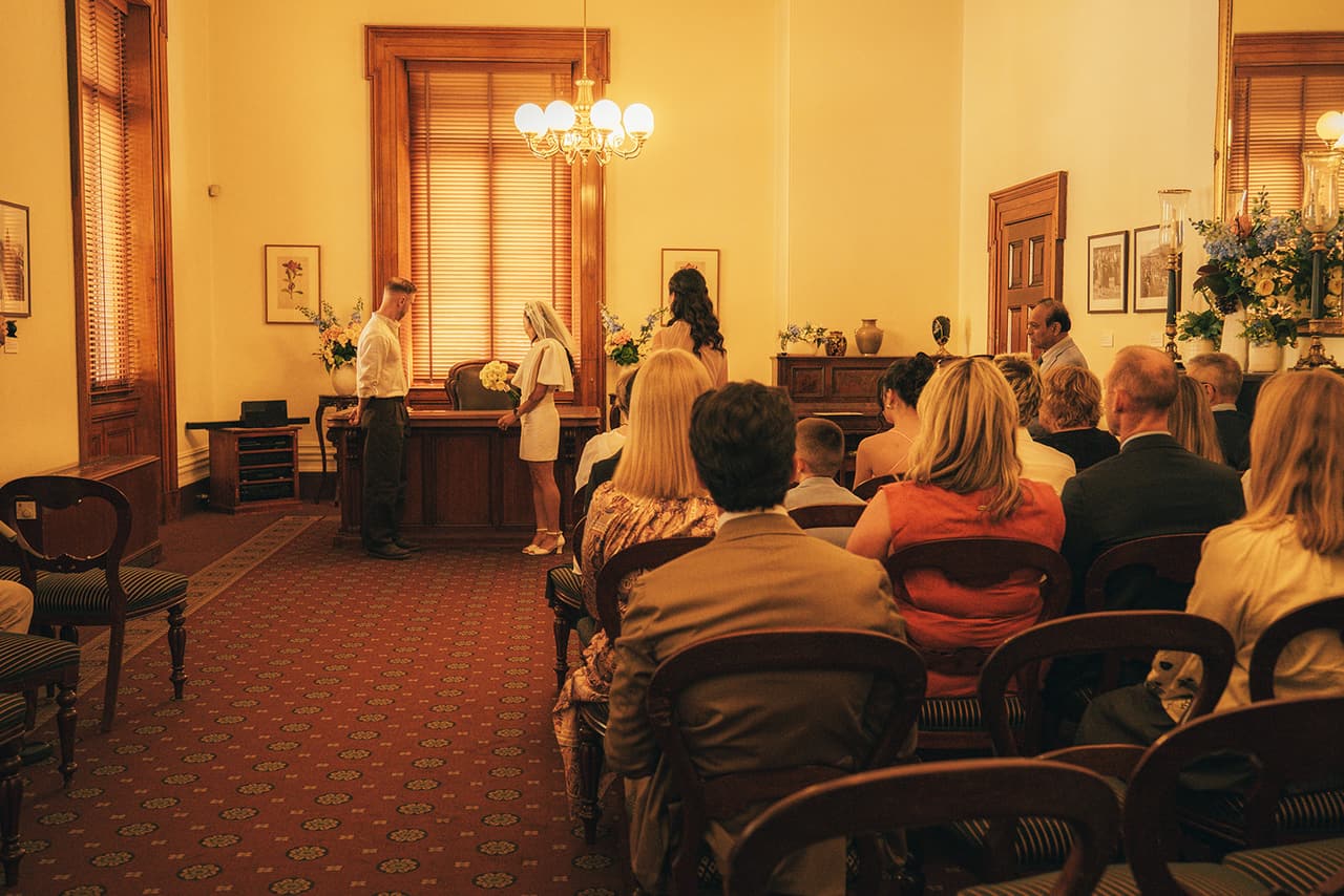 The Margaret Craig Room full of guests, seen from behind the chairs, with Cecile and Stefan at the registry desk under the tall window and chandelier