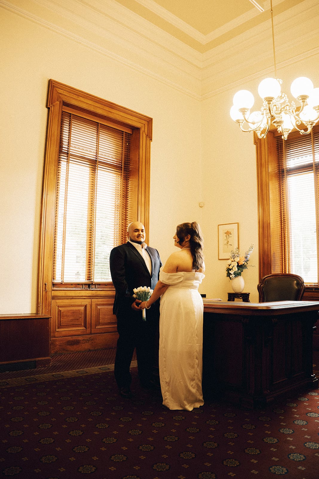 Martina and Diego at the registry desk in the corner of the ceremony room, two tall windows with timber blinds and a chandelier above