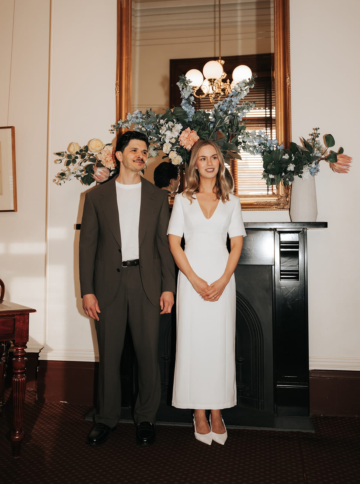 Karina and Chris in front of the black fireplace of the small ceremony room, a gilded mirror, a chandelier and blue and peach flowers above the mantel