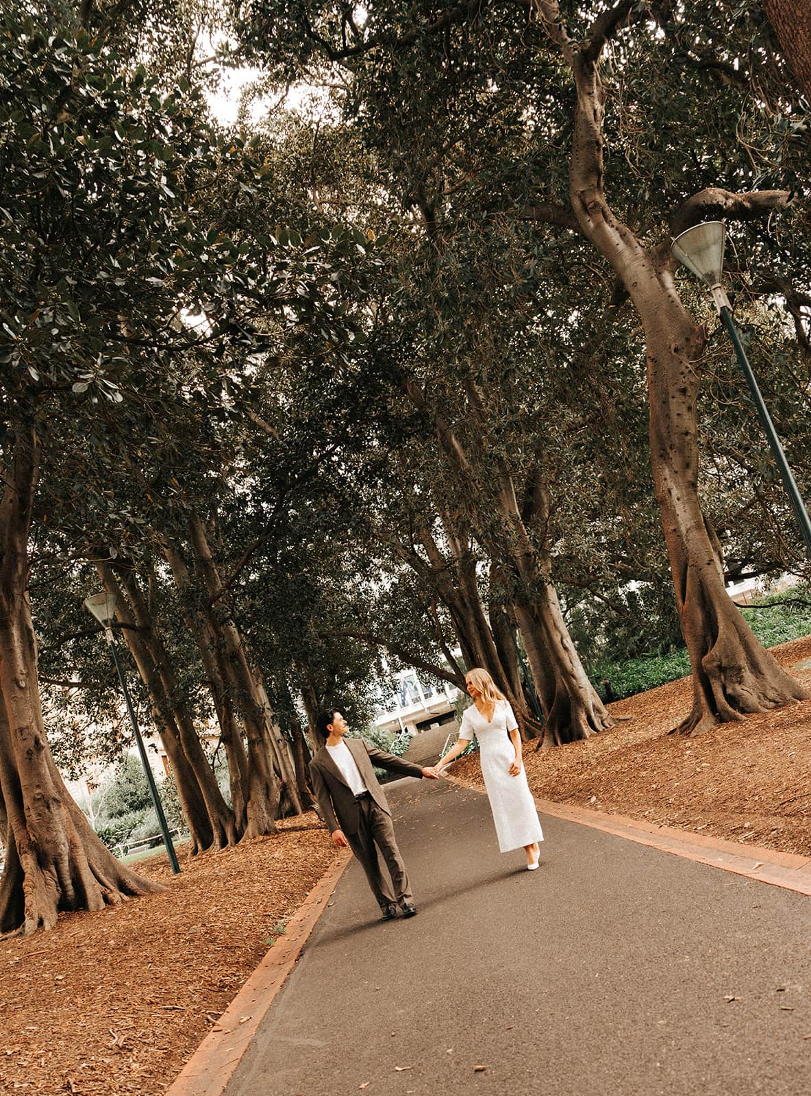Karina and Chris walking hand in hand along a curving path under the fig trees of Treasury Gardens