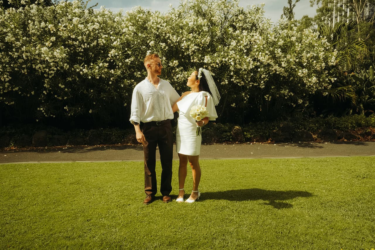 Cecile and Stefan on a sunny lawn in front of a tall hedge of white flowers in the gardens beside the Old Treasury Building