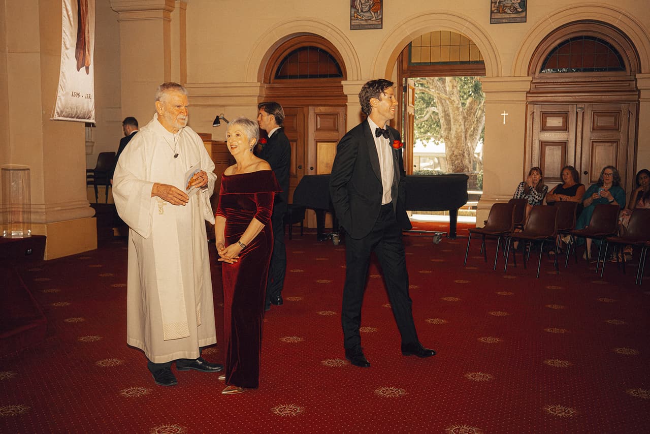 The priest with a guest in burgundy velvet and a man in black tie at the front of the chapel before the ceremony, arched doorways open behind them