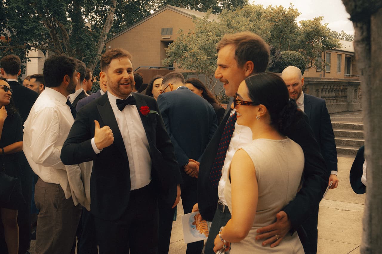 Guests talking on the terrace outside the chapel after the ceremony, a stone balustrade and trees behind them