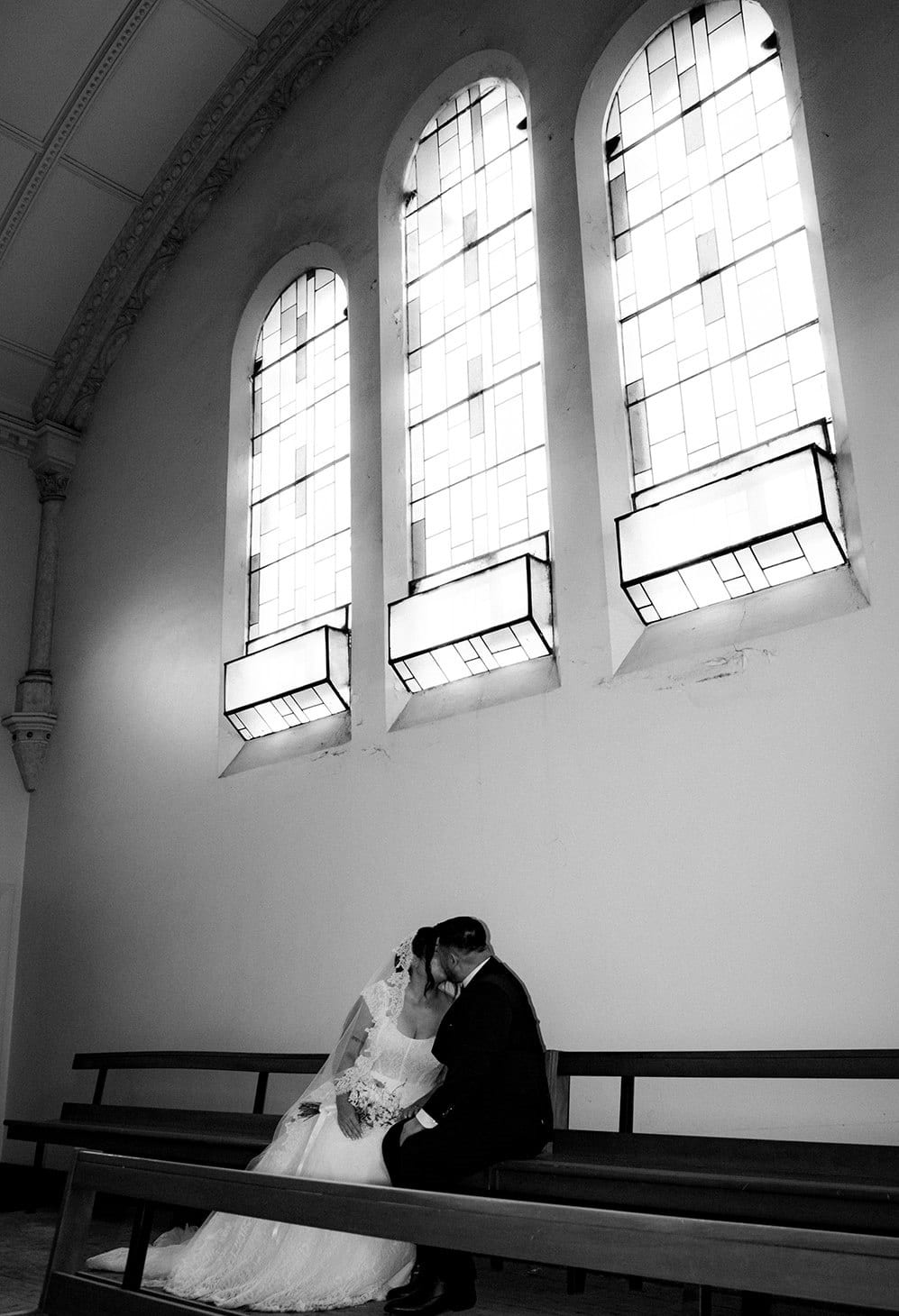 Black and white photo of Ashley and Bryan sitting close on a pew beneath three tall windows