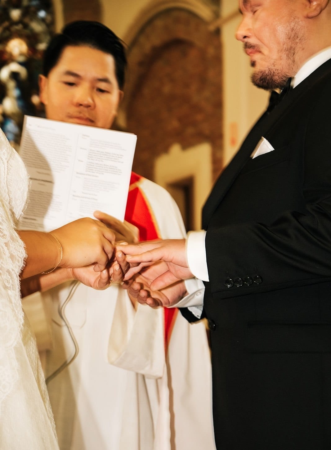 Bryan placing the ring on Ashley’s finger while the priest reads from the booklet
