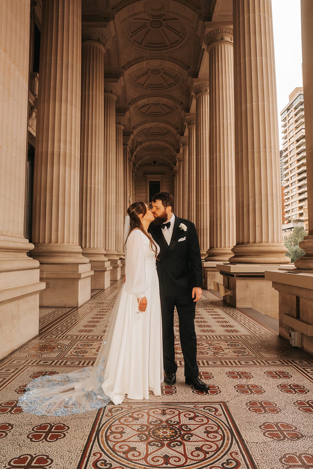 Bianca and Aidan kissing in the long colonnade of Parliament House, its patterned tiles and vaulted ceiling running away behind them