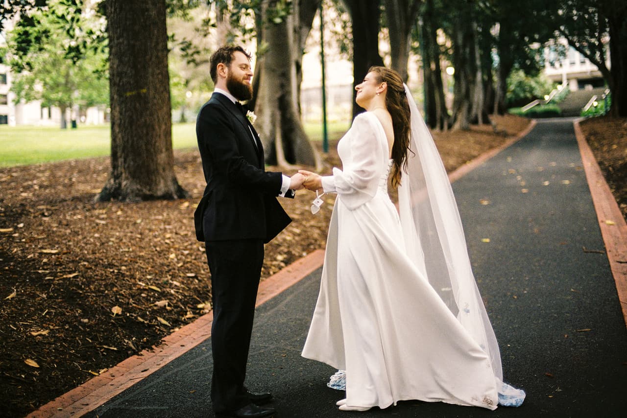 Bianca and Aidan holding hands on the avenue path, her veil trailing behind her, on 35mm film