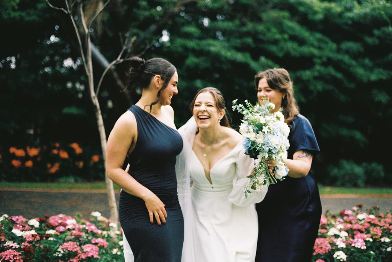 Bianca laughing between two friends in navy dresses, her blue and white bouquet in hand, on 35mm film