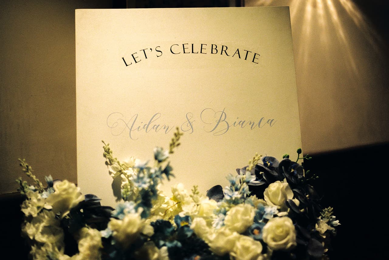 A sign reading Let’s Celebrate with Aidan and Bianca’s first names above white and blue flowers, on 35mm film