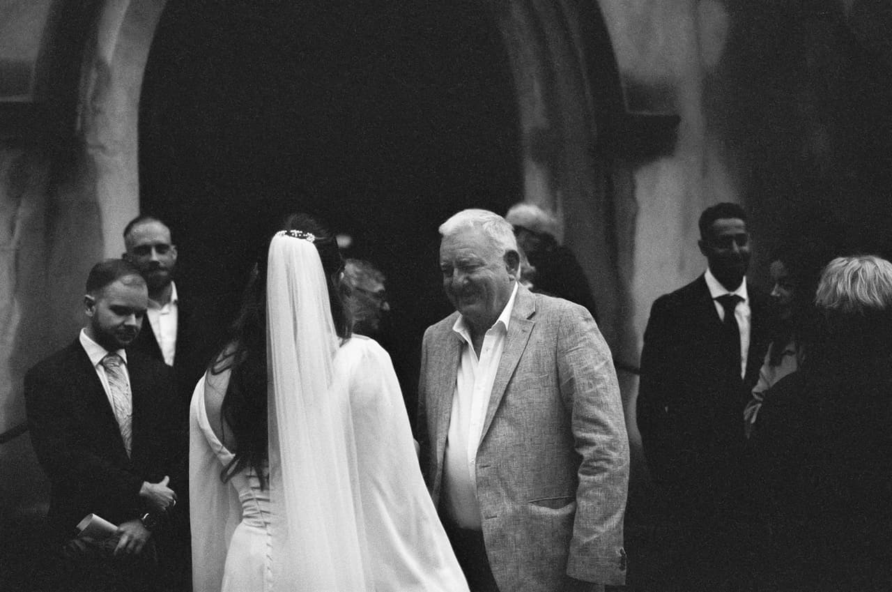 Black and white photo of Bianca, her veil down her back, greeting smiling guests in the church porch, on 35mm film