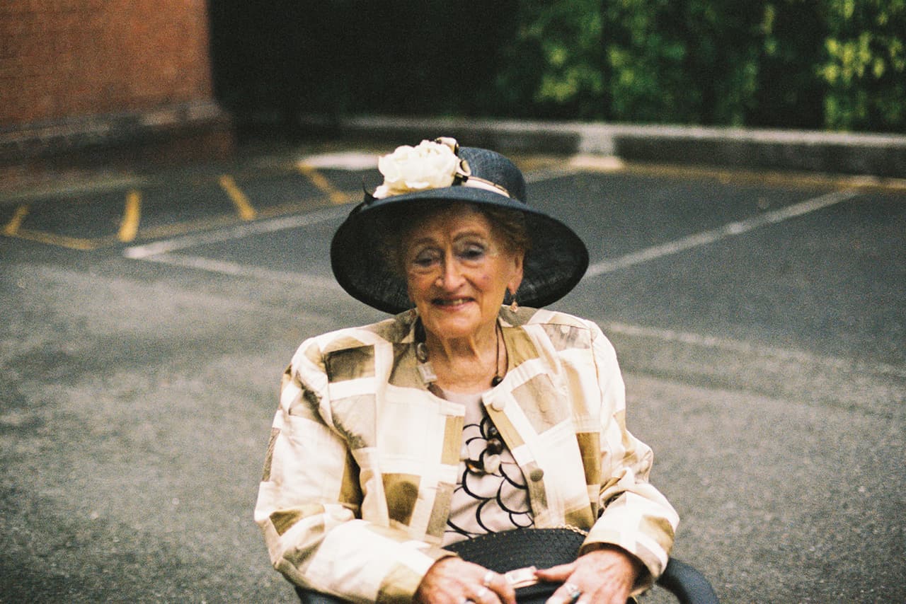 An older guest in a dark hat with a cream flower and a patterned jacket, smiling as she sits outside the church, on 35mm film
