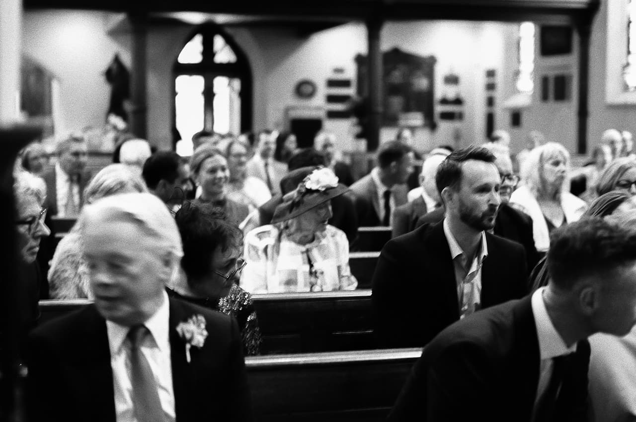 Black and white photo of guests in the pews during the ceremony, an older guest in a flowered hat among them, on 35mm film