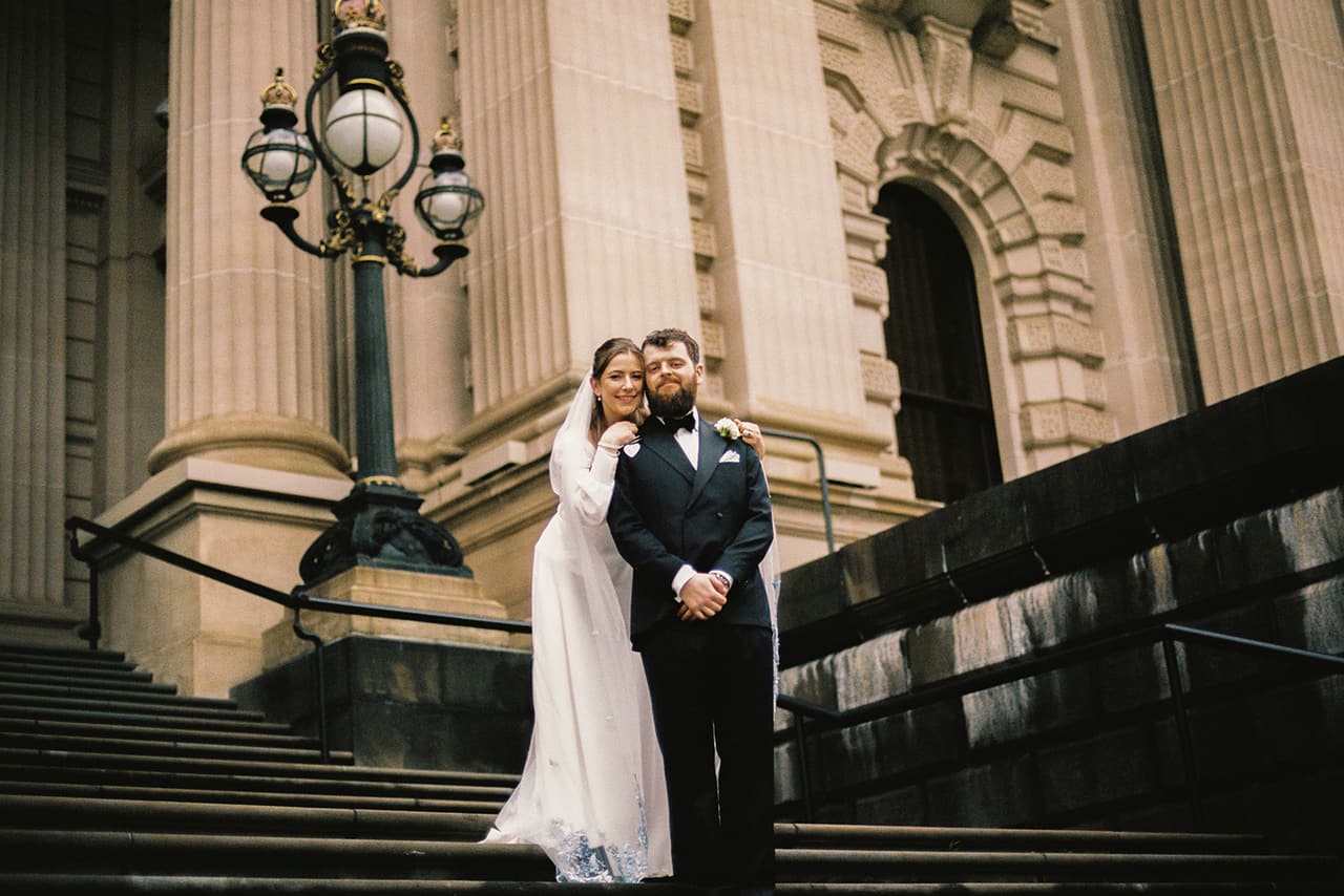 Bianca and Aidan standing together on the steps of Parliament House beside an old lamp, on 35mm film
