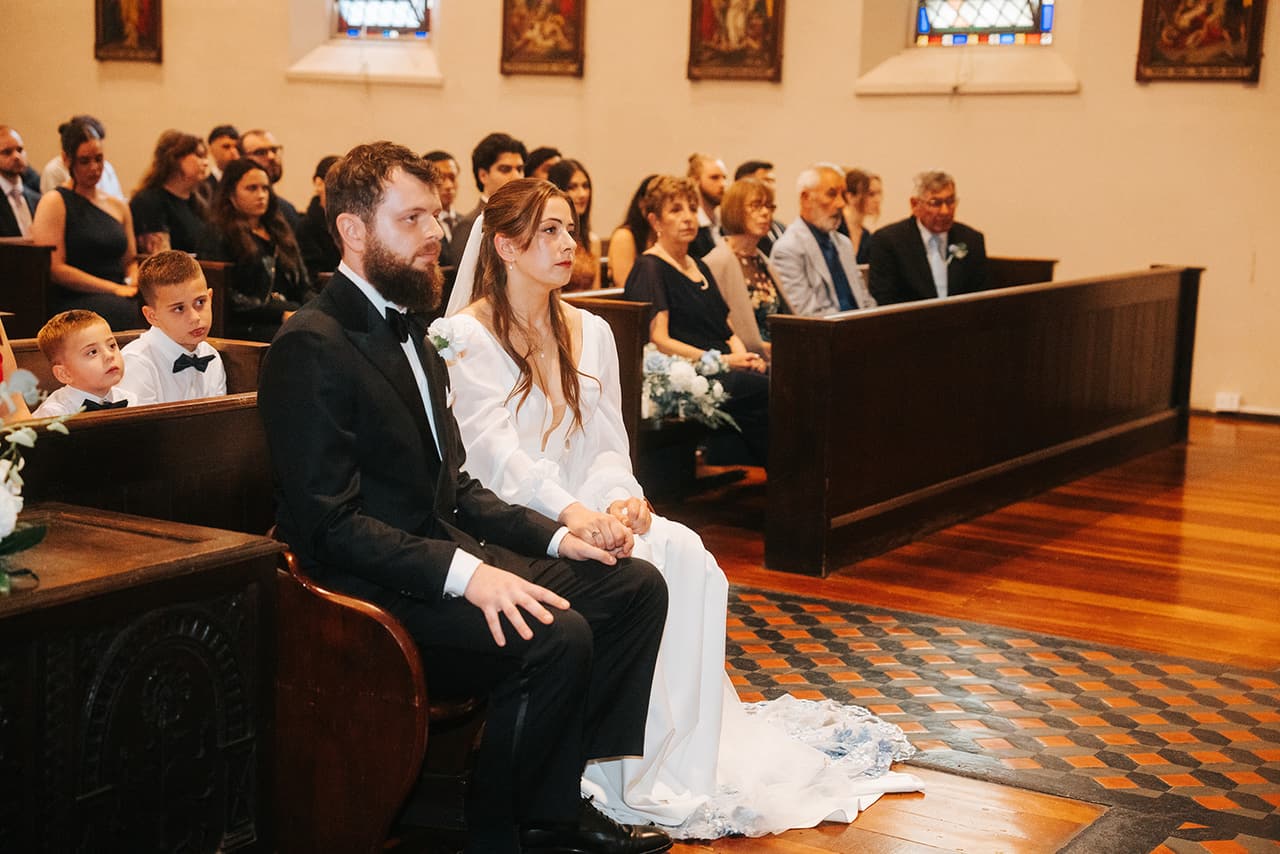 Bianca and Aidan holding hands as they sit in the front pew, guests in the rows behind them