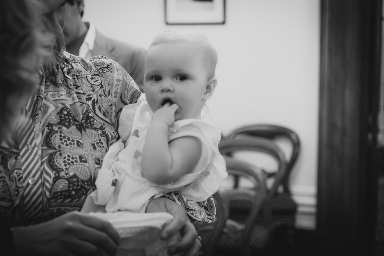 Black and white photo of a baby chewing a hand in a guest’s arms in the ceremony room