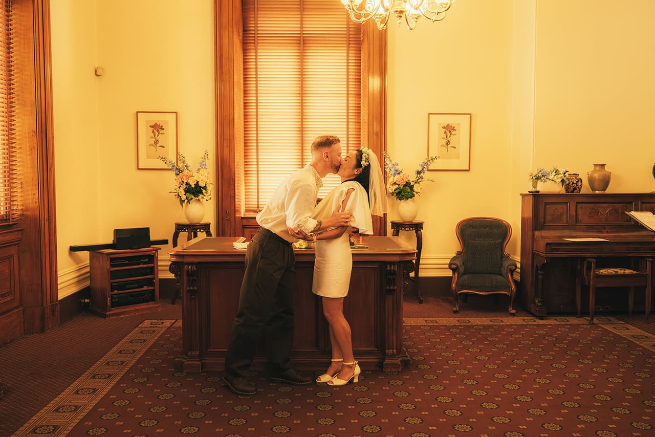 Cecile and Stefan kissing at the registry desk under the chandelier, framed botanical prints on the walls