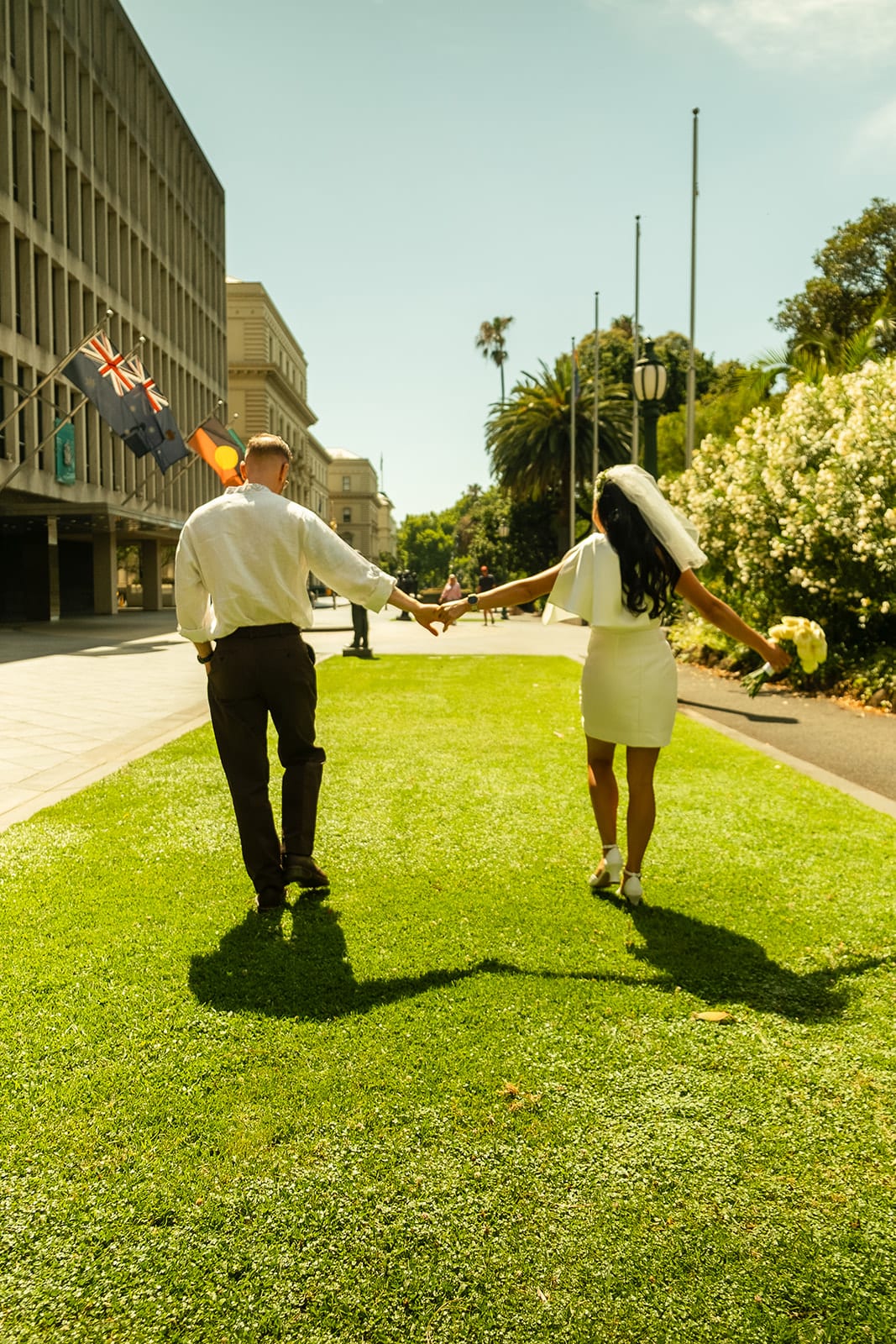 Cecile and Stefan walking away hand in hand down a sunny strip of lawn, flags flying on the building beside them